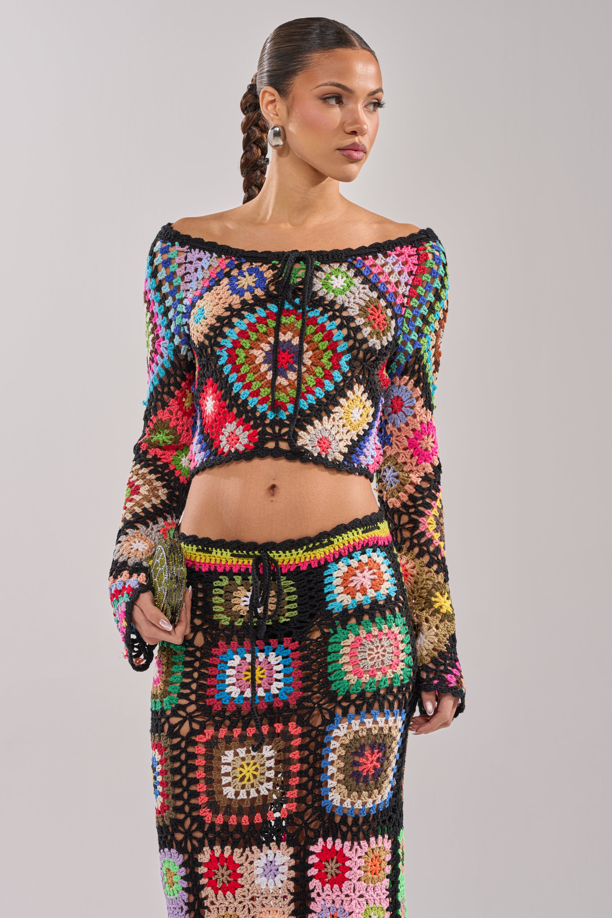 A woman with braided hair wears the SHINE ON YOU CROCHET TOP and matching skirt in multicolored granny-square patchwork, creating a festival-ready look. She holds a small clutch and stands against a light gray background. 1