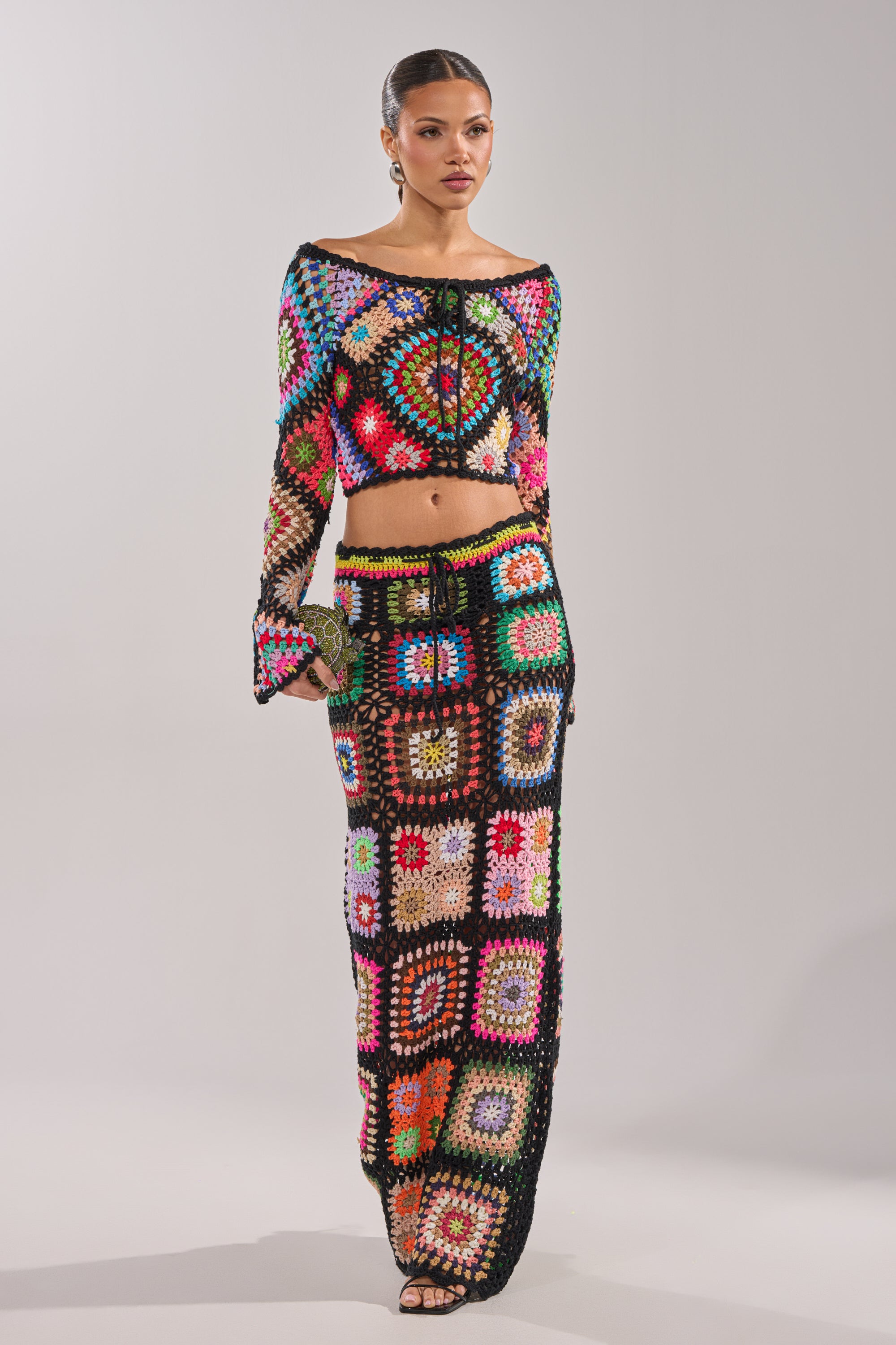 A woman wears the SHINE ON YOU CROCHET TOP with a matching long skirt, creating a vibrant, festival-ready look while holding a small clutch purse against a plain background. 7