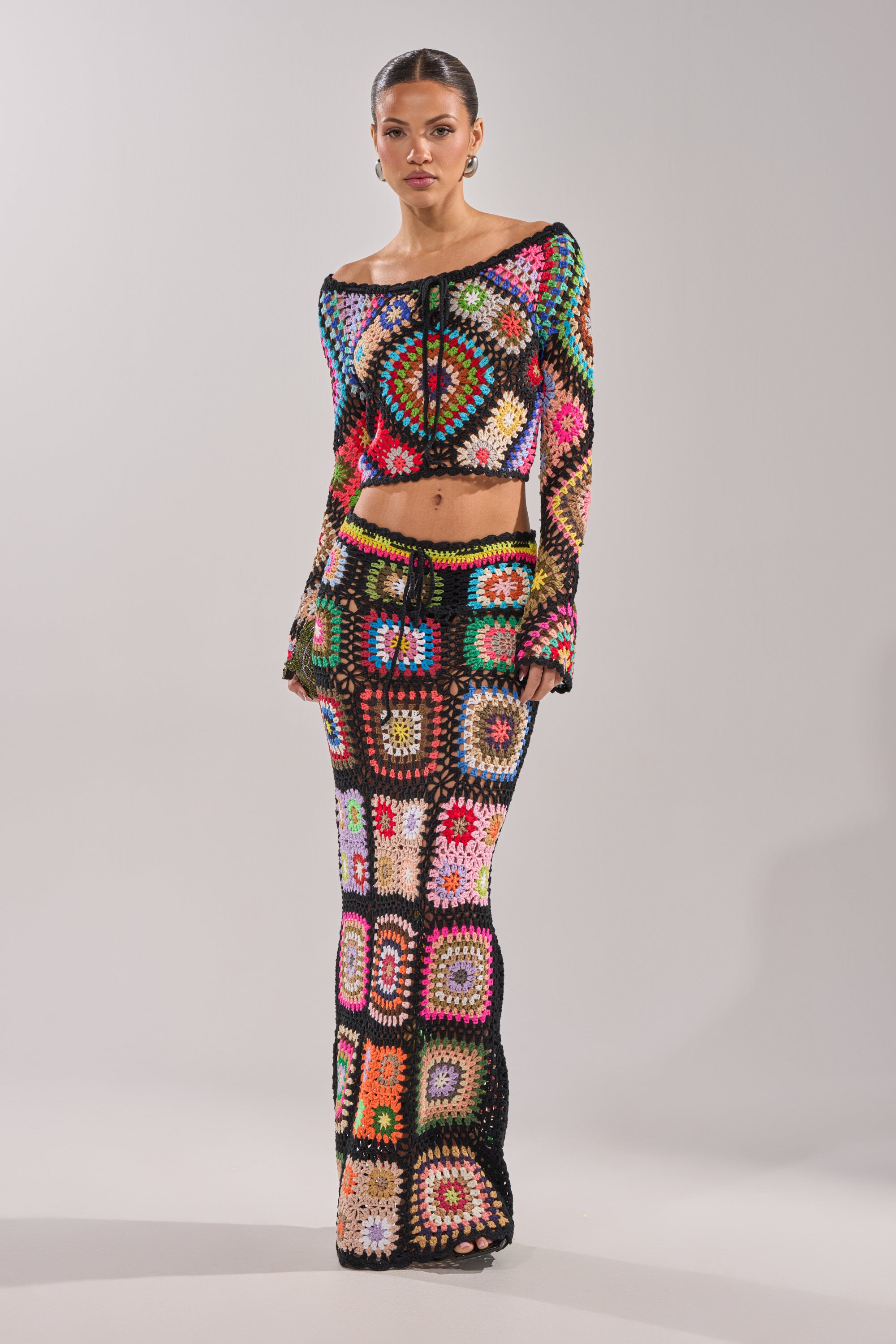 A woman poses against a plain light background wearing the SHINE ON YOU CROCHET TOP, a colorful, long-sleeve patchwork crop top with multicolored squares and a fitted, festival-ready design. 8