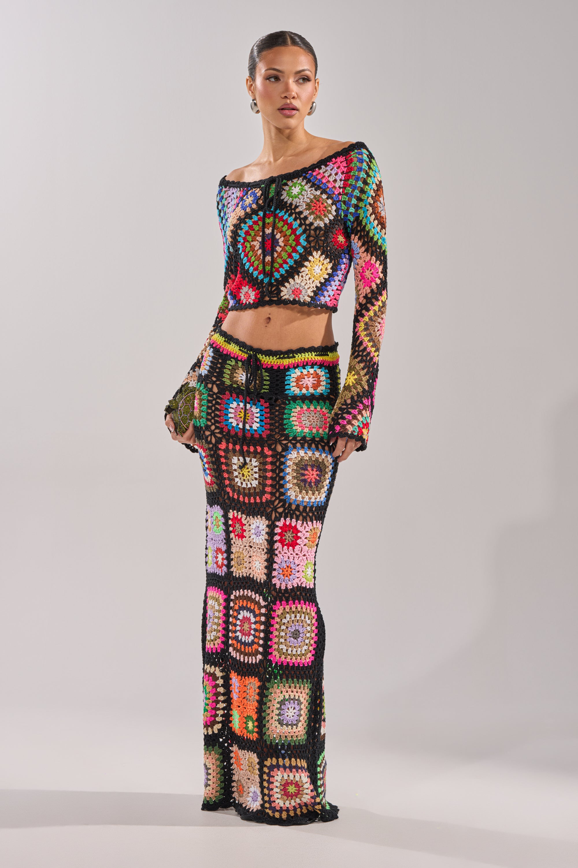 A woman models the SHINE ON YOU CROCHET MAXI SKIRT, a colorful high-rise crochet maxi skirt with a matching granny-square long-sleeve crop top, both showcased against a plain light gray background. 0