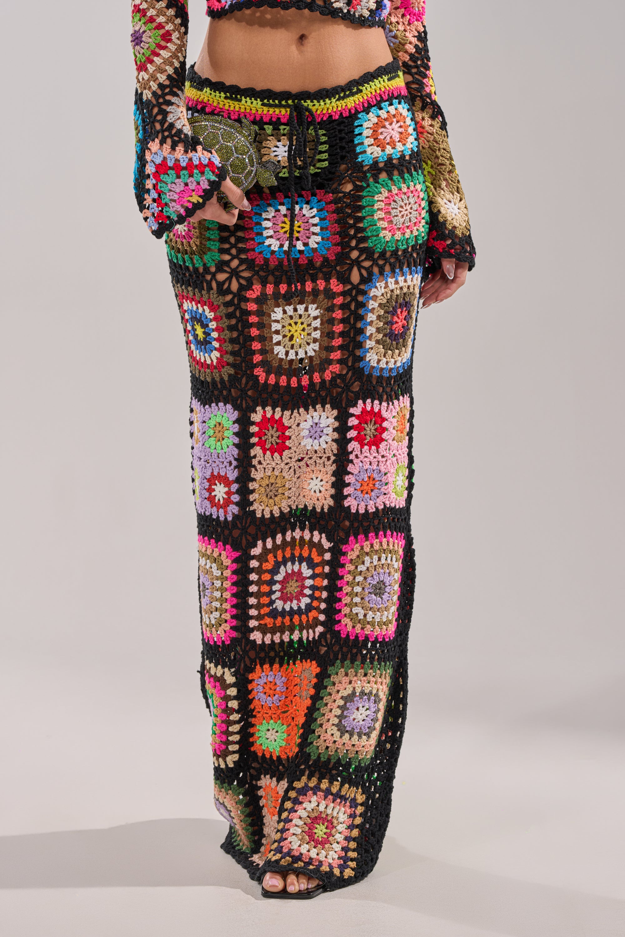 The lower half of a person is shown wearing the SHINE ON YOU CROCHET MAXI SKIRT, featuring vibrant granny-square patchwork and a high-rise waist, while holding a small green clutch bag. 2