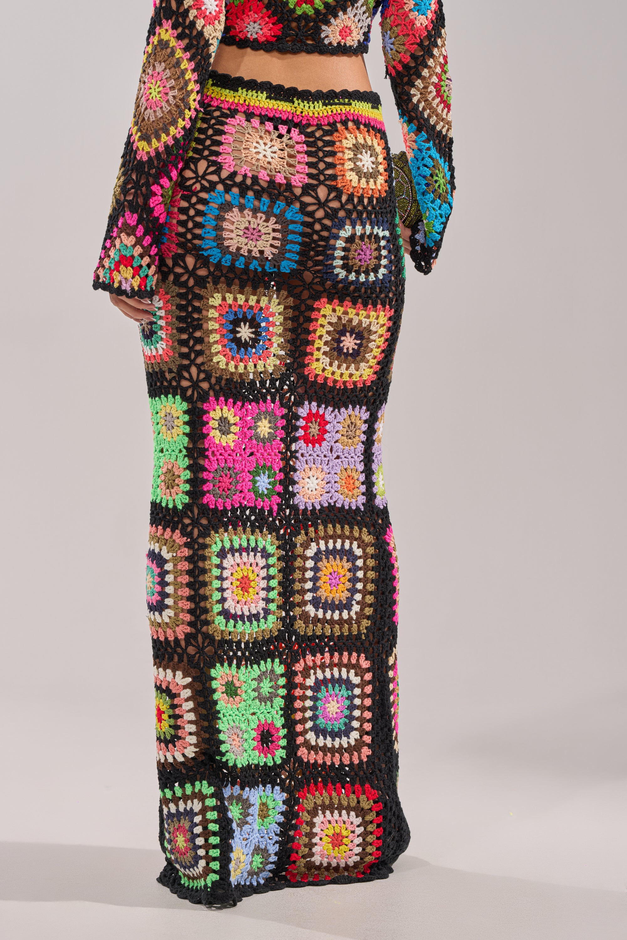 Someone stands against a plain light background wearing the SHINE ON YOU CROCHET MAXI SKIRT, a high-rise, black-based maxi skirt with multicolored granny-square patchwork, paired with a matching crochet top. 5