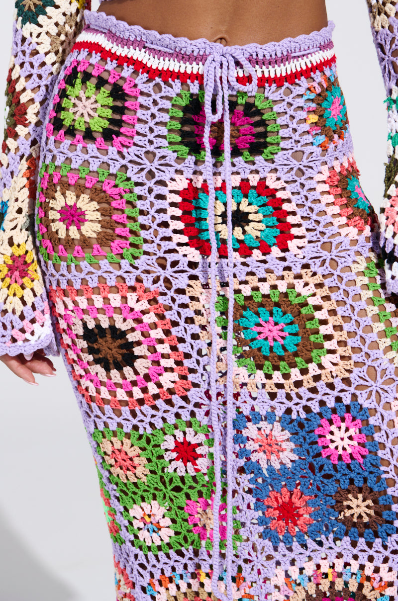 The SHINE ON YOUR CROCHET MAXI SKIRT is a lively crochet maxi skirt with multicolored granny squares and a lilac waistband, ideal for summer. The high-rise fit includes a string tie, covering the lower body with intricate patterns. A hand holds the skirt's left side gently.