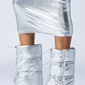 A person wearing a shiny metallic faux leather skirt and the AZALEA WANG DHRUV SILVER PUFFER BOOTIE, featuring zippers and ties. The mid-calf shaft enhances its futuristic allure, complemented by a sleek stiletto heel. The outfit stands out against a plain, light-colored background.