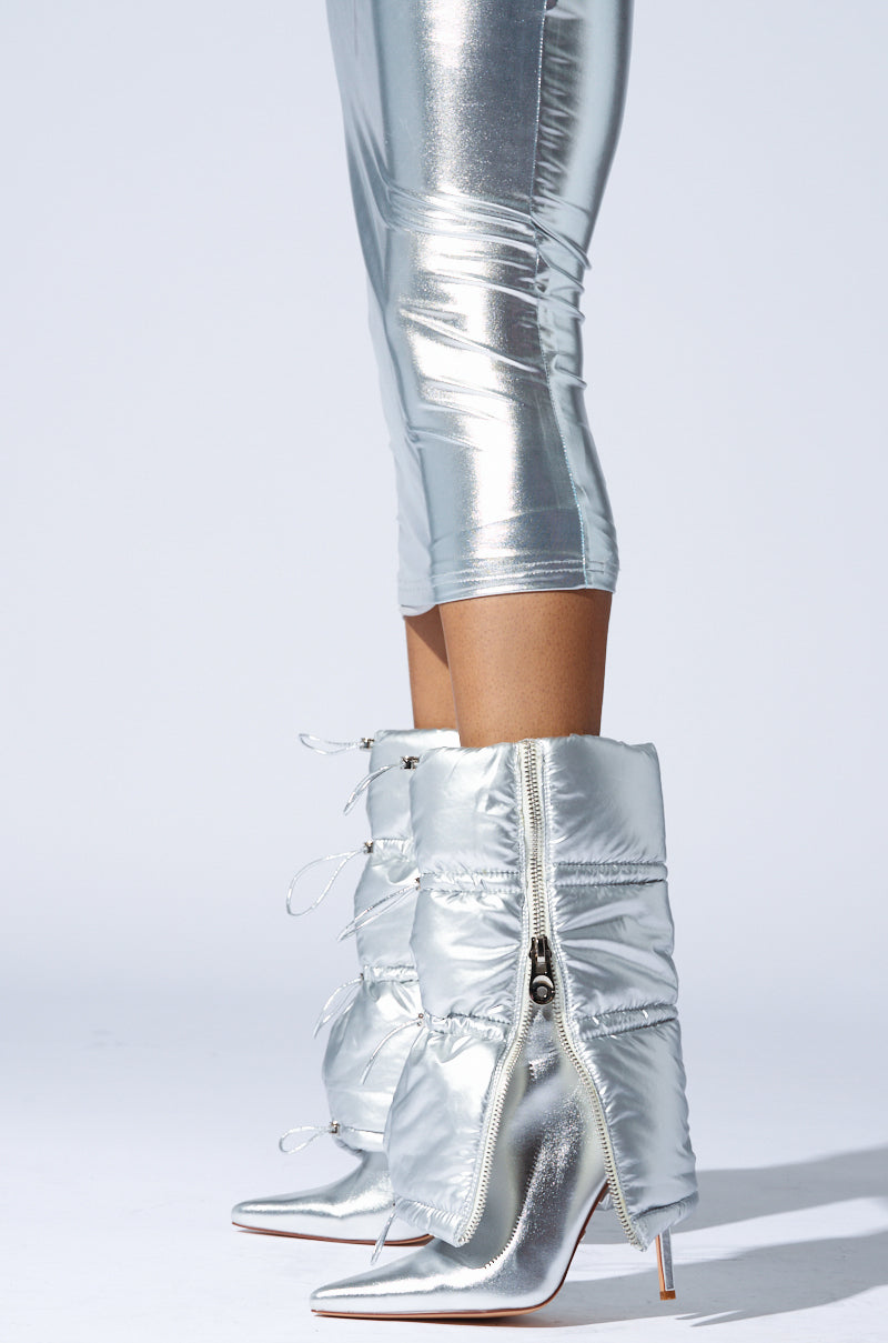 A person is wearing shiny silver metallic faux leather leggings paired with the AZALEA WANG DHRUV SILVER PUFFER BOOTIE, which features zipper detailing and an exaggerated, puffy design. The background is plain white.