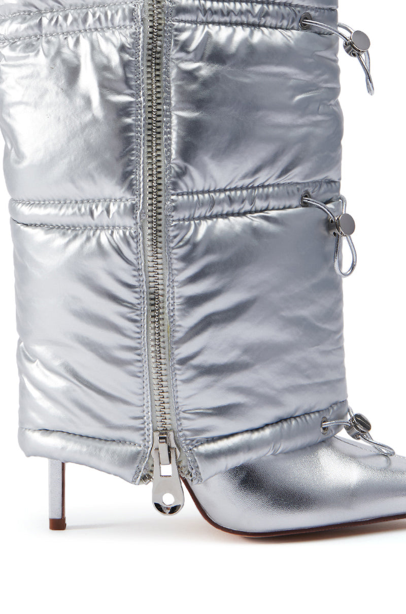 The AZALEA WANG DHRUV SILVER PUFFER BOOTIE is shown in a close-up view, highlighting its metallic faux leather texture and high-heeled design. This silver puffer ankle boot features a prominent front zipper, drawstring details with metal toggles, and a slender stiletto heel. The shot focuses on the sleek side profile of the boot.