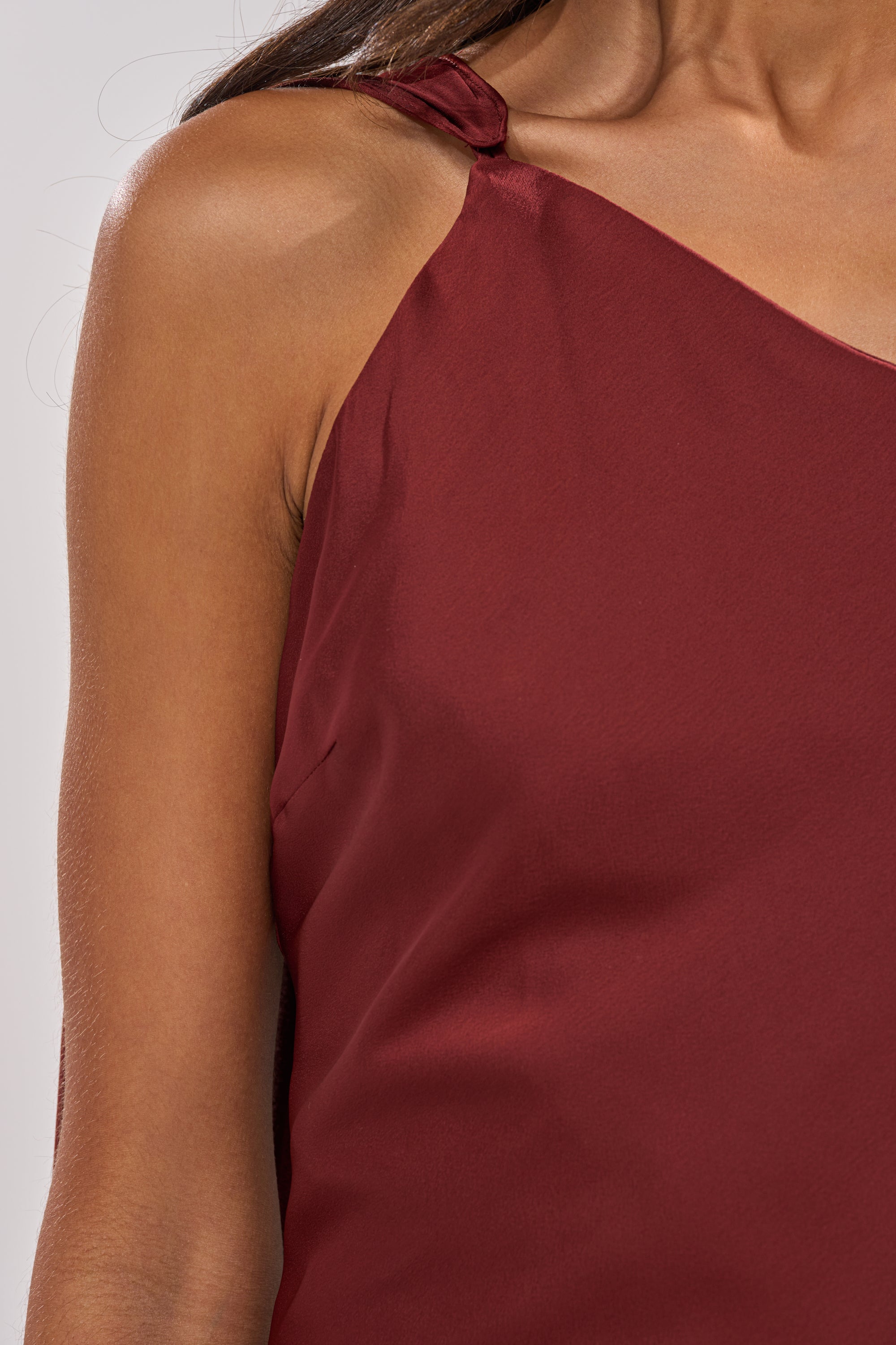 A close-up of a woman's shoulder and upper chest wearing the RSVP READY SATIN MAXI DRESS, featuring a sleeveless, dark red satin design with an asymmetrical neckline, set against a plain light background. 9
