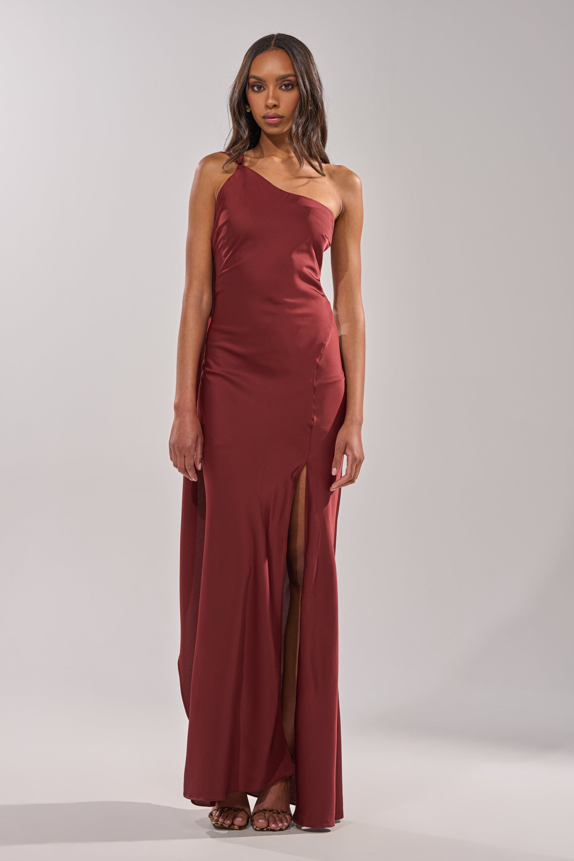 A woman models the RSVP READY SATIN MAXI DRESS, which has a sleeveless design, asymmetrical neckline, and high side slit. She poses in sandals with long, wavy hair against a plain light gray backdrop. 1