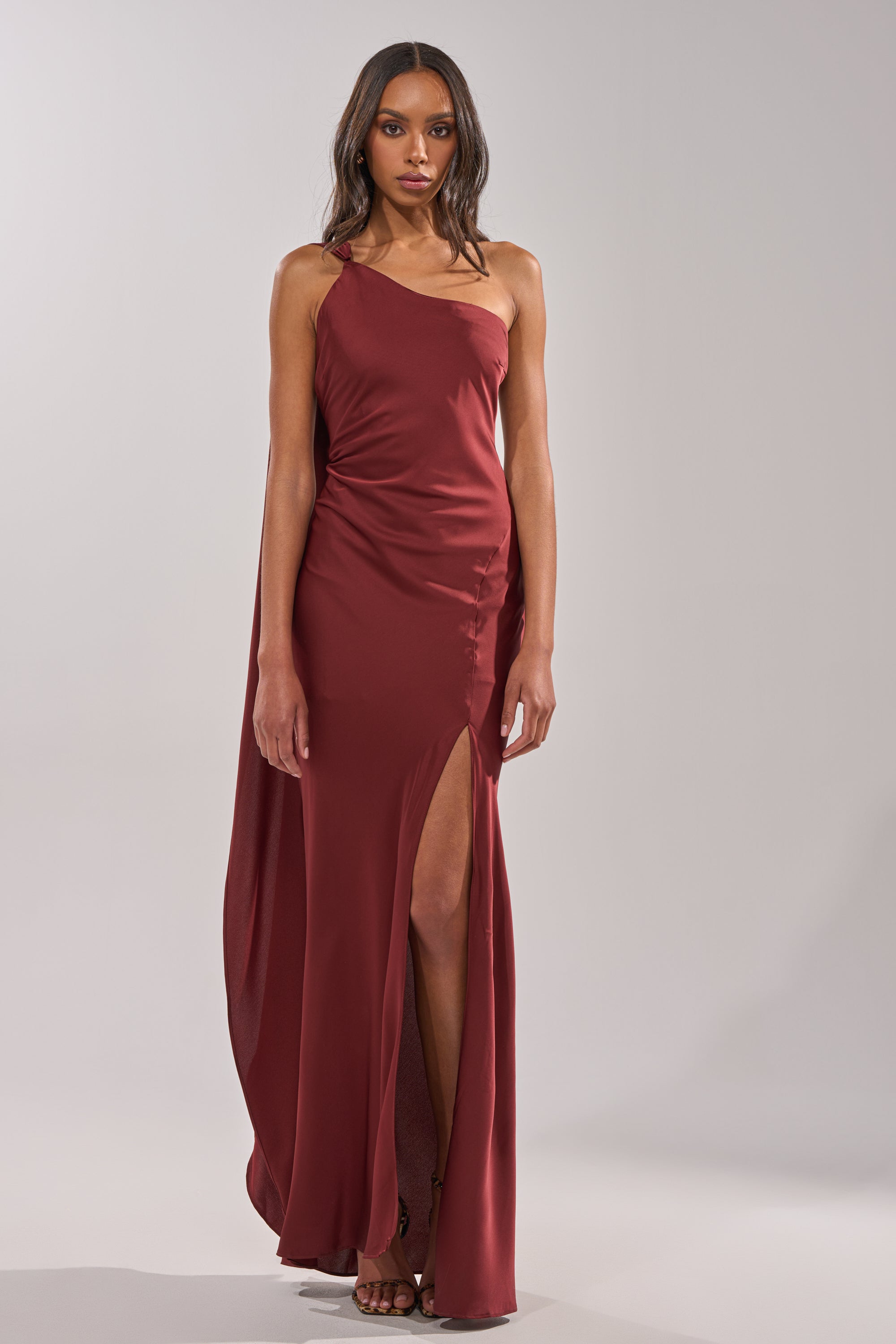 A woman poses in the RSVP READY SATIN MAXI DRESS, a sleeveless maroon maxi with a side slit, paired with strappy heels, against a plain light gray background. 2