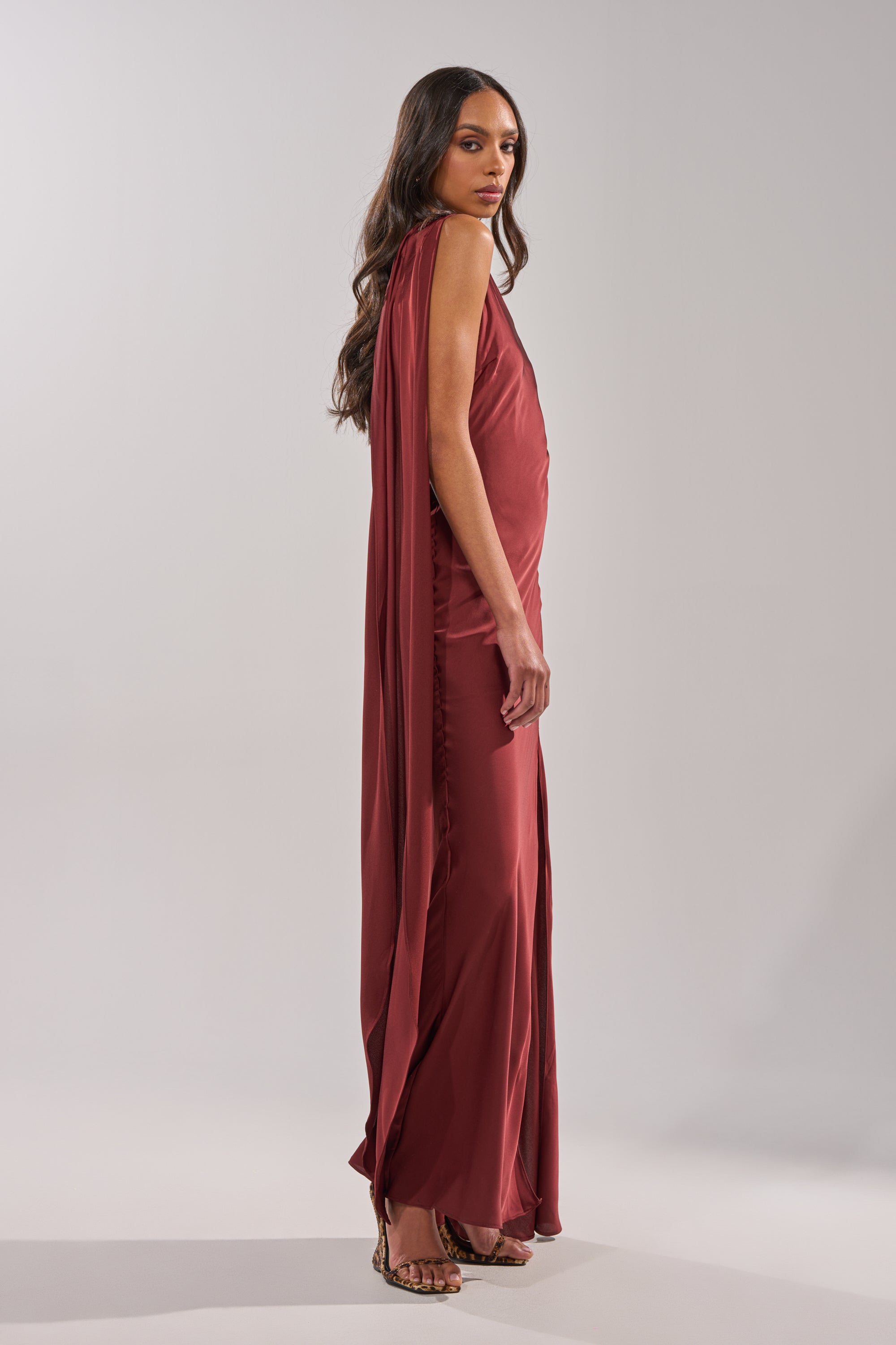 A woman with long wavy hair stands sideways in the RSVP READY SATIN MAXI DRESS, a floor-length, dark red gown featuring an asymmetrical neckline and draped back detail, paired with strappy heels against a plain light background. 4