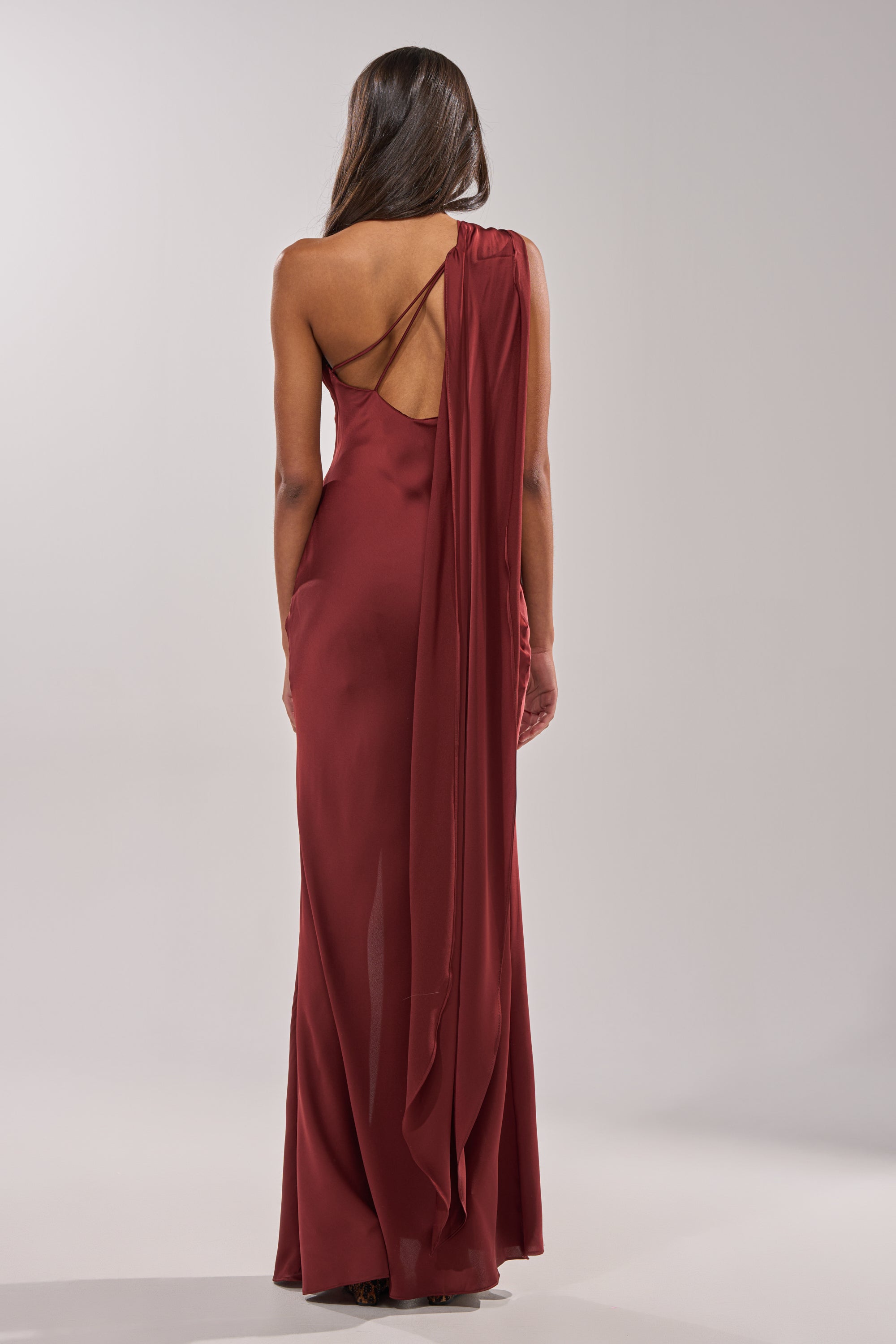 A woman with long brown hair models the RSVP READY SATIN MAXI DRESS, a floor-length, sleeveless burgundy gown with an asymmetrical neckline and draped back, standing against a plain light background. 5