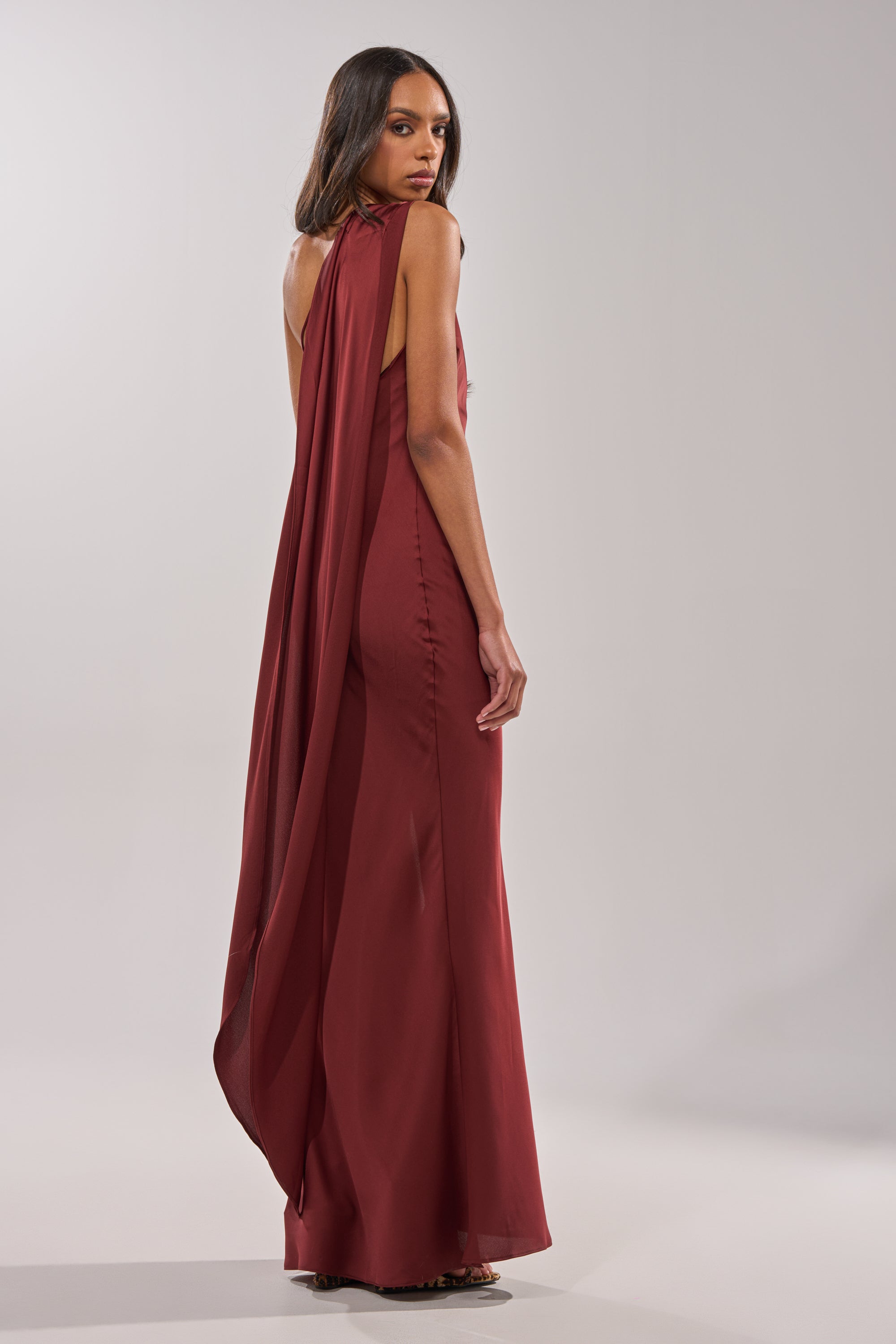 A woman poses against a plain light gray background, wearing the RSVP READY SATIN MAXI DRESS—a sleeveless, floor-length burgundy gown with a flowing cape detail draped from her shoulders. 6