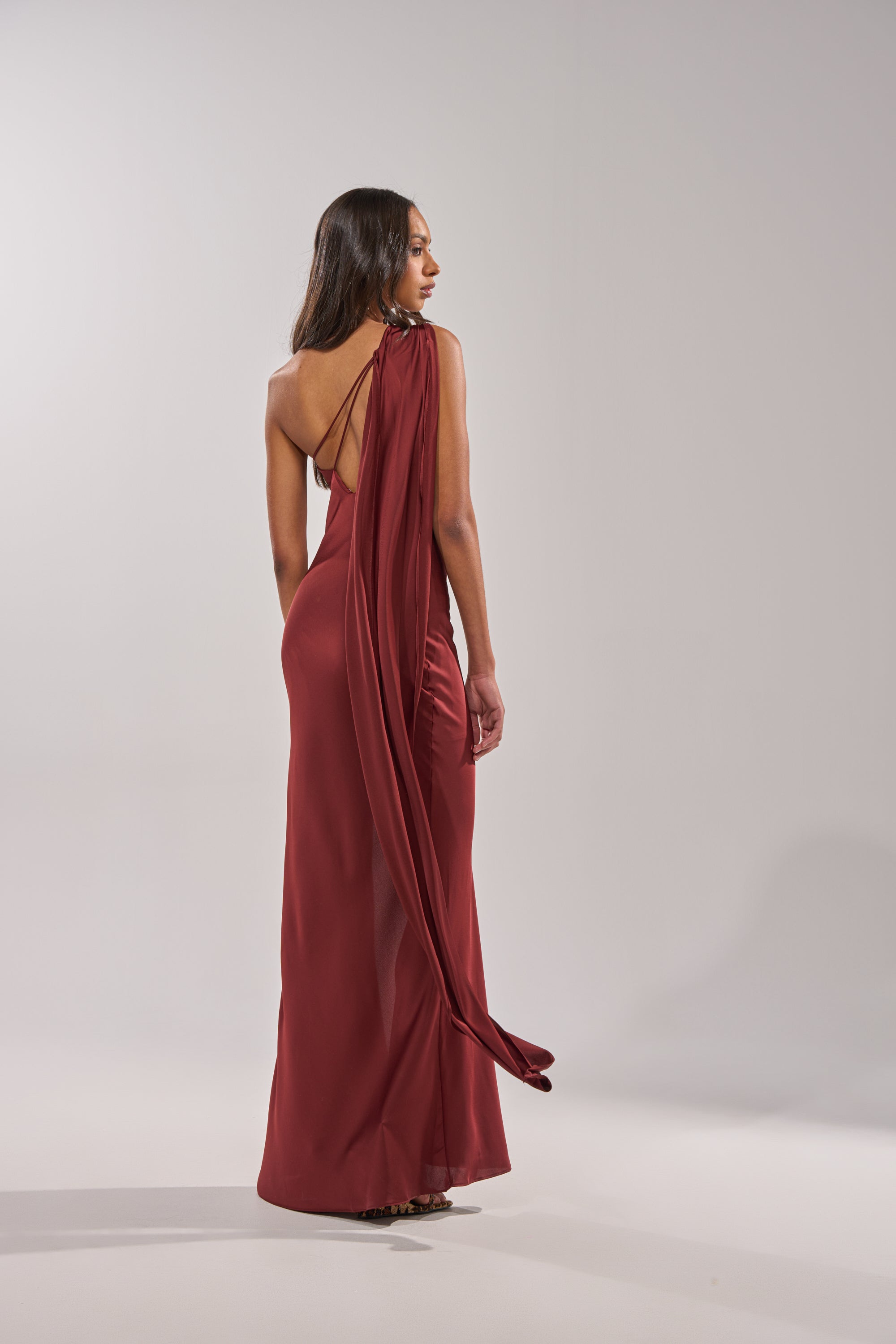 A woman in the RSVP READY SATIN MAXI DRESS—floor-length, sleeveless satin with an open back and a flowing sash—stands against a plain light background, looking over her shoulder. 7