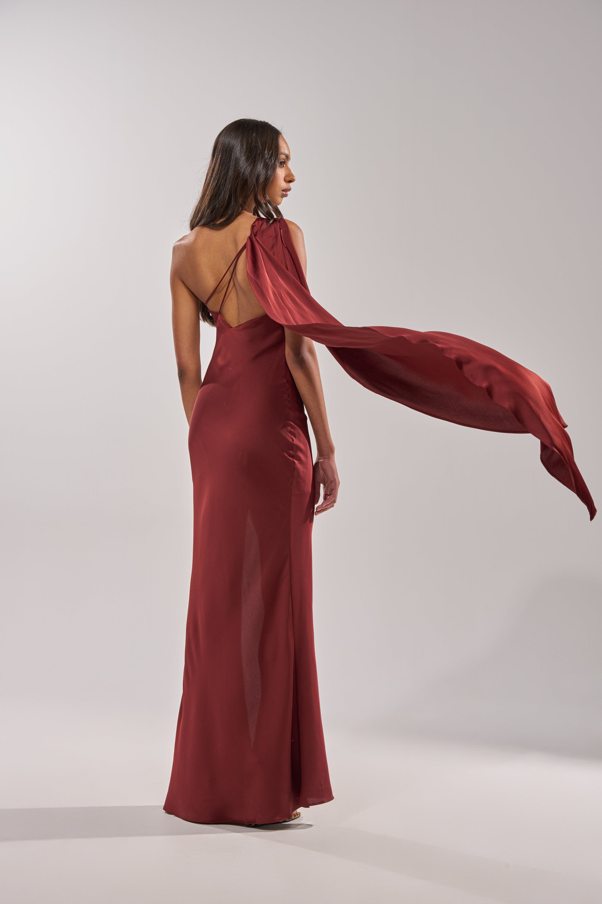 A woman faces away wearing the RSVP READY SATIN MAXI DRESS, a floor-length dark red gown with an asymmetrical neckline and flowing shoulder detail. She stands against a softly lit, plain background. 8