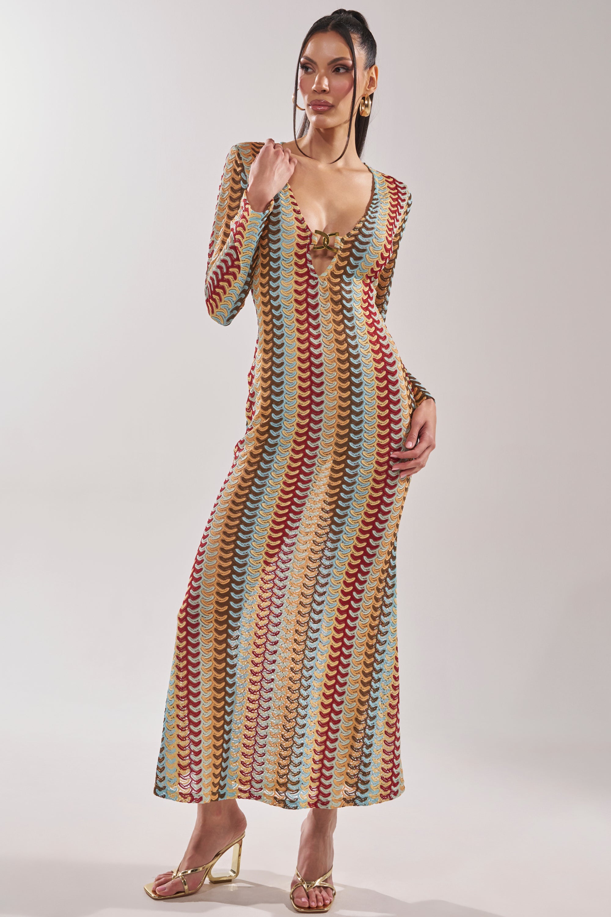 A woman wears the THREAD THEORY CROCHET MAXI DRESS, a long, fitted multicolor knit with a deep V-neck, long sleeves, and wavy red, yellow, teal, and beige stripes. She pairs it with gold heels and matching earrings. 2