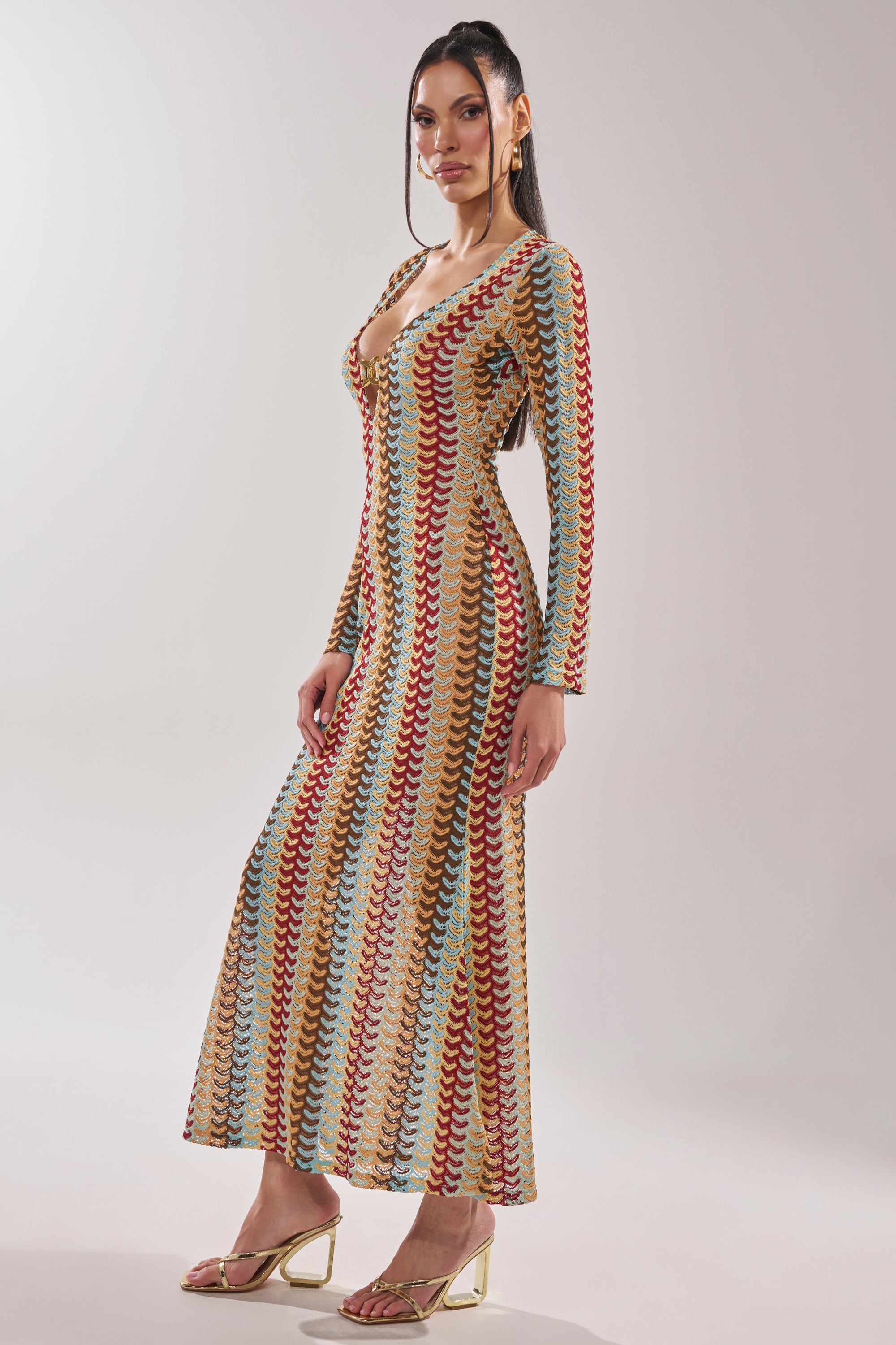A woman poses against a plain, light background in the THREAD THEORY CROCHET MAXI DRESS, a long, form-fitting multicolor knit dress with a deep neckline and wavy stripes. She pairs it with strappy gold heels and has straight, dark hair. 3