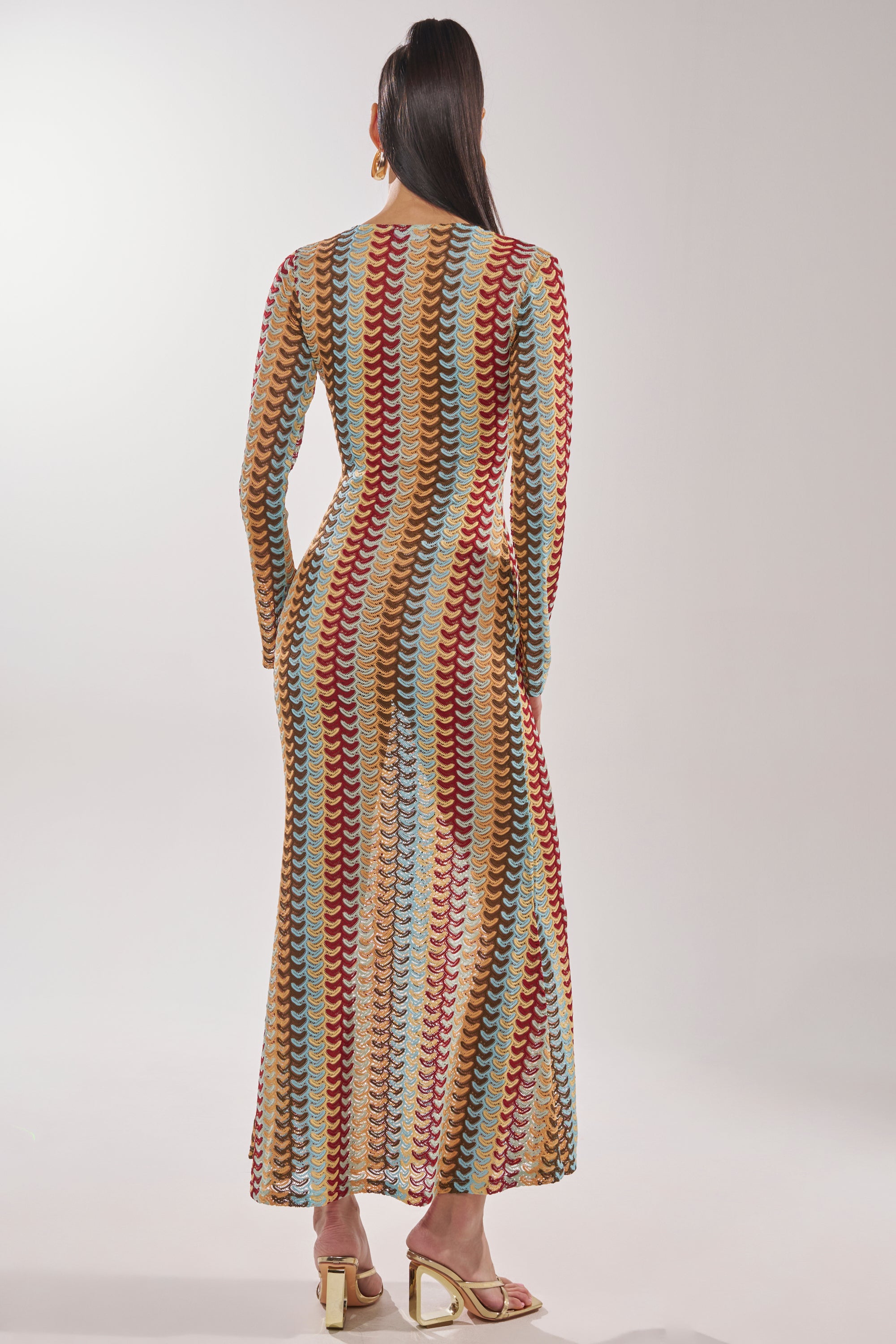 A person stands facing away, wearing the THREAD THEORY CROCHET MAXI DRESS with long sleeves and a multicolor wavy zigzag crochet pattern, paired with gold heeled sandals against a plain light background. 4