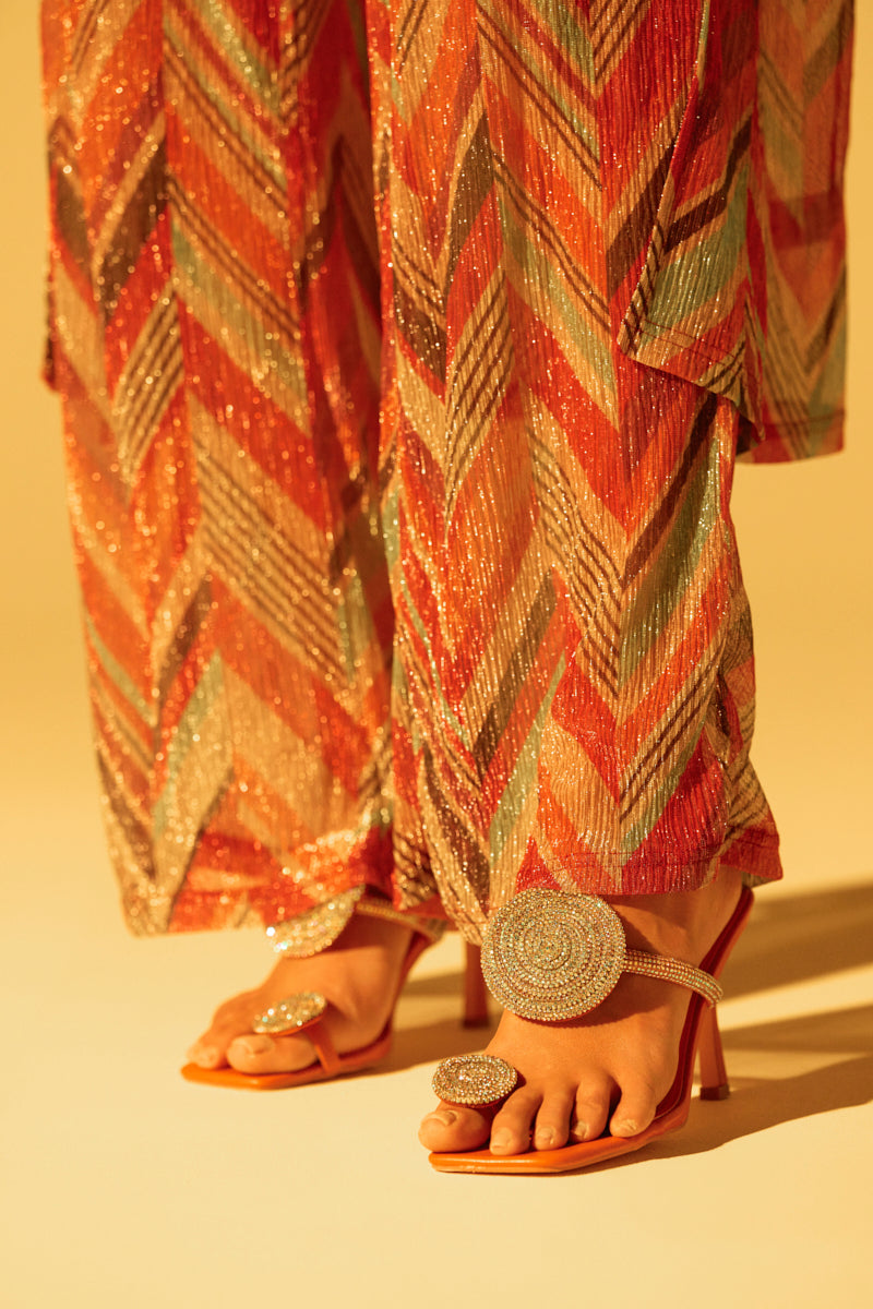 A person is wearing patterned pants with zigzag designs in orange, pink, and green, paired with AZALEA WANG DIARA beige raffia embellished stiletto sandals. The background is a warm, soft yellow.