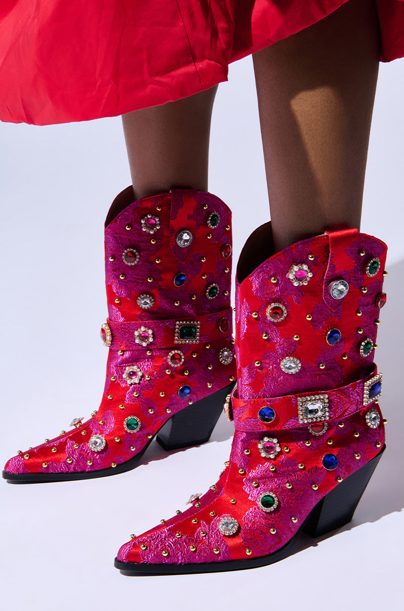 A person wearing the AZALEA WANG DILIGENT RED BROCADE AND GEMS WESTERN BOOTIE, adorned with colorful rhinestones and intricate embroidery, stands on a white background. The bottom edge of a red skirt is visible, perfectly complementing the bootie's vibrant design.