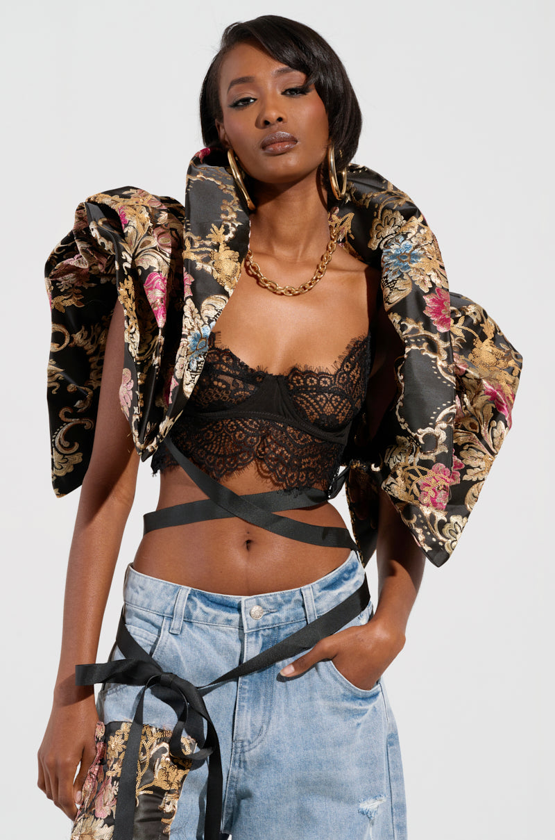 A person poses confidently, wearing the DION STATEMENT BOLERO IN BROCADE with voluminous sleeves, a black lace crop top, and light blue jeans. They accessorize with large hoop earrings and a chunky gold chain necklace.