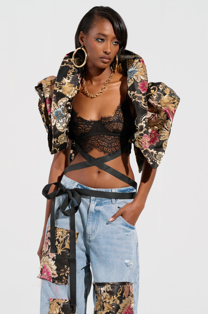 A fashionable person wearing the Dion Statement Bolero in brocade, a black lace top, and denim pants with floral patches poses confidently. They accessorize with large hoop earrings and a gold necklace, one hand resting casually in their pocket.