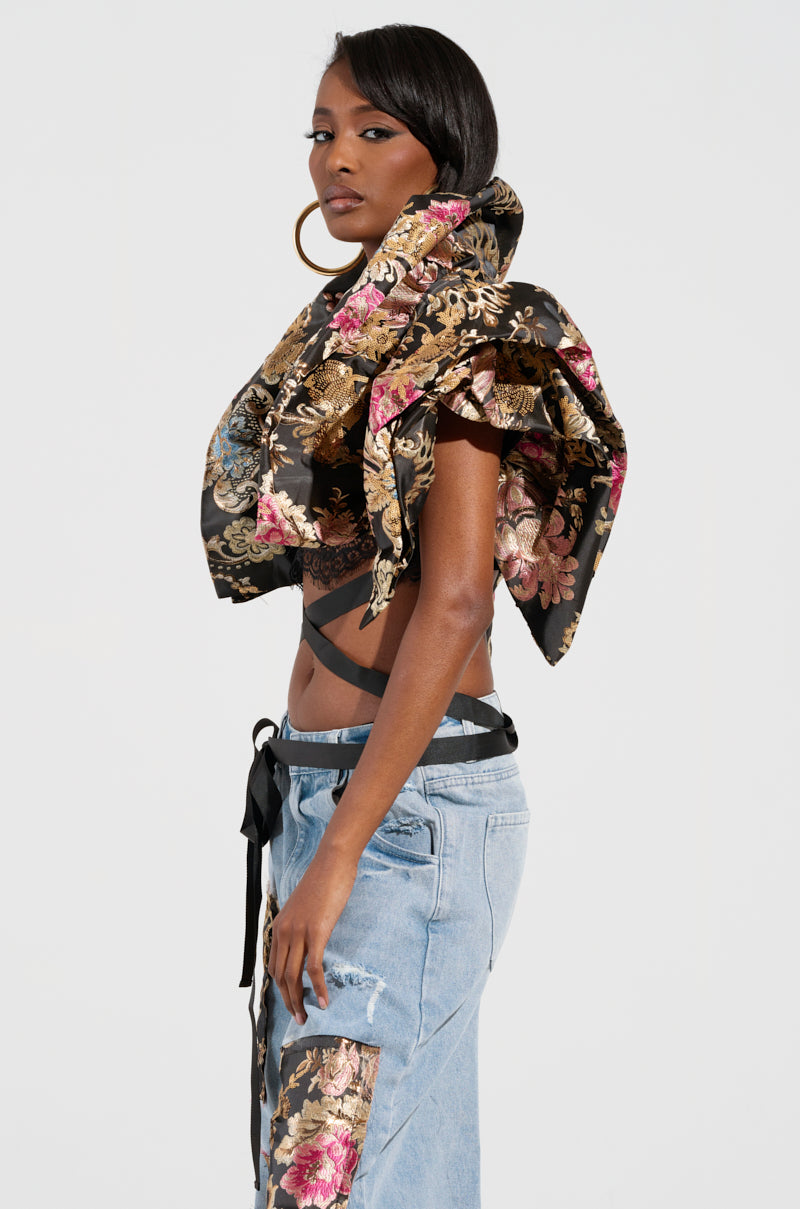 A woman poses confidently in a stylish outfit featuring a floral, ruffled top with an asymmetrical design and distressed denim pants adorned with floral patches. She accessorizes with large hoop earrings and completes the look with the elegant Dion Statement Bolero in Brocade.