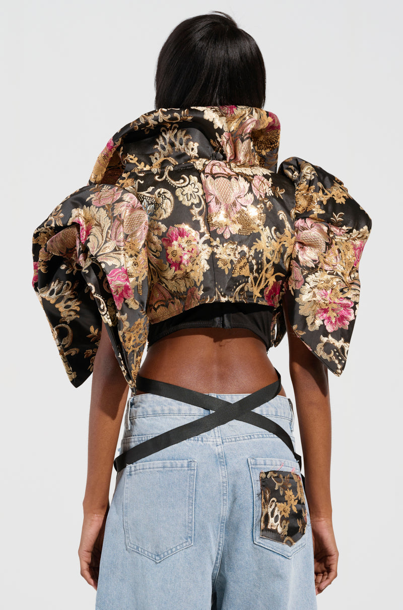 A person stands facing away from the camera, wearing the DION Statement Bolero in black and gold brocade with exaggerated shoulders. They pair this striking jacket with light blue jeans that highlight a matching floral pocket and black crisscross straps.