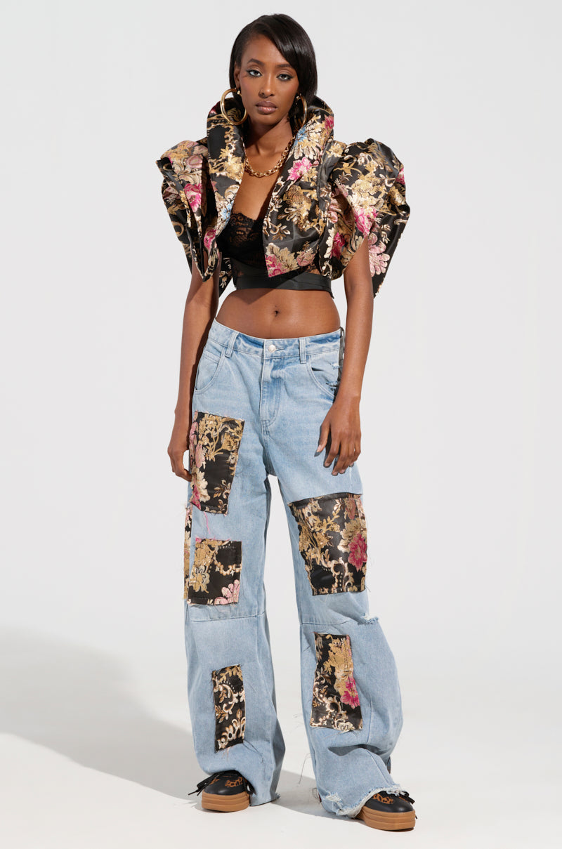 A person wearing the DION Statement Bolero in Brocade, an exquisite floral-patterned top with dramatic ruffled shoulders, paired with patchwork jeans adorned with matching floral fabric and subtle sequin embellishments. The outfit masterfully fuses modern and baroque elements, with the individual posing confidently on a plain white background.