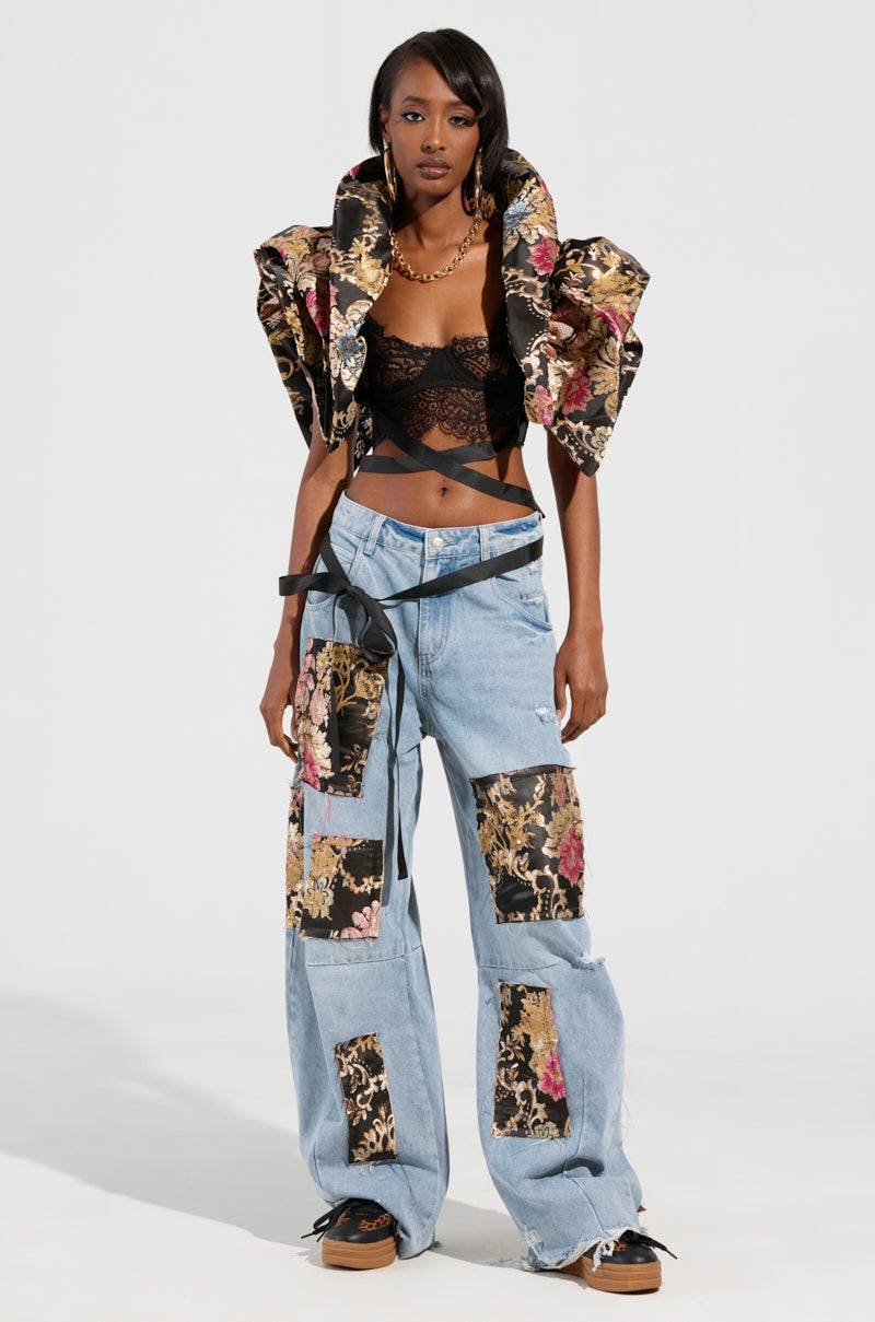 A woman stands confidently against a light background, wearing a bold outfit with a black lace top, the eye-catching Dion Statement Bolero in Brocade, and loose blue jeans featuring floral patchwork.