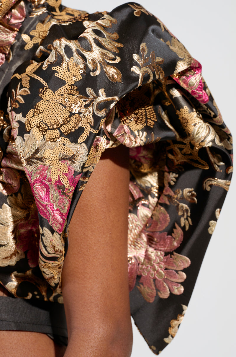 A close-up view of a person wearing the DION STATEMENT BOLERO IN BROCADE, showcasing its intricate floral pattern in gold, pink, and beige on a shiny black fabric. The bolero features ruffled designs and is embellished with sequins, creating a dramatic and elegant appearance.