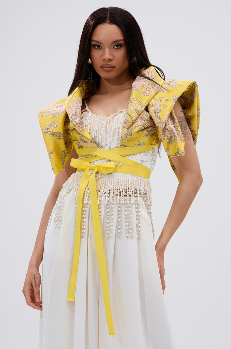 A woman stands against a plain light background wearing a detailed white dress with pearl accents and sculptural shoulders, styled with the DION STATEMENT BOLERO IN YELLOW tied at the waist.