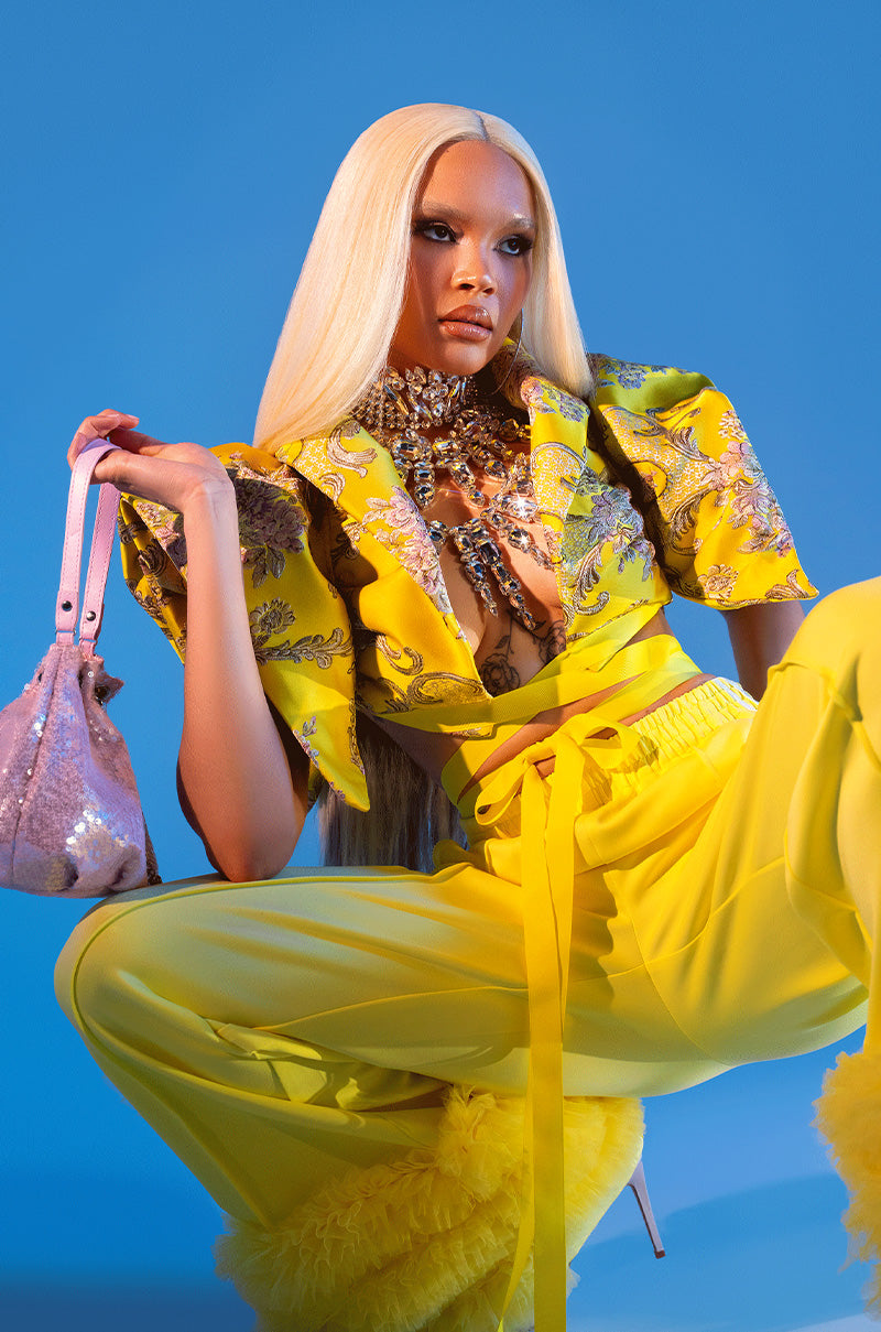 A woman with long, straight platinum blonde hair poses confidently in the DION STATEMENT BOLERO IN YELLOW and wide-leg ruffled pants, holding a sparkling pink handbag and wearing bold jewelry against a vivid blue background.