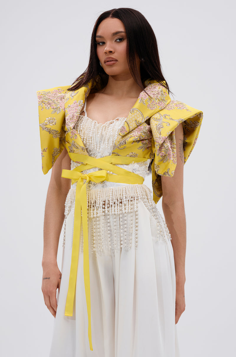 A woman wears the DION STATEMENT BOLERO IN YELLOW, featuring sculptural shoulders and floral jacquard, over a pearl-embellished white top and flowing white pants, standing against a plain light background.
