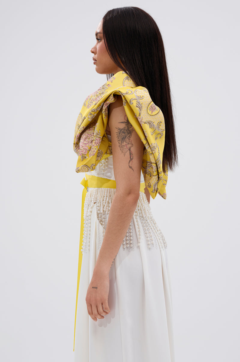 A woman in profile stands against a plain background, wearing the DION STATEMENT BOLERO IN YELLOW over a white dress with pearl details. She has straight dark hair and a visible floral tattoo on her upper arm.
