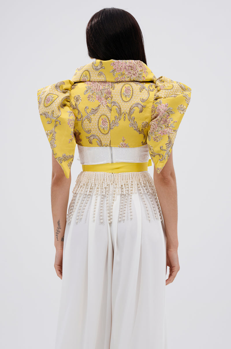 A woman stands facing away, wearing the DION STATEMENT BOLERO IN YELLOW with sculptural shoulders and a floral jacquard pattern, paired with a long, flowing white skirt adorned with intricate bead detailing at the waist.