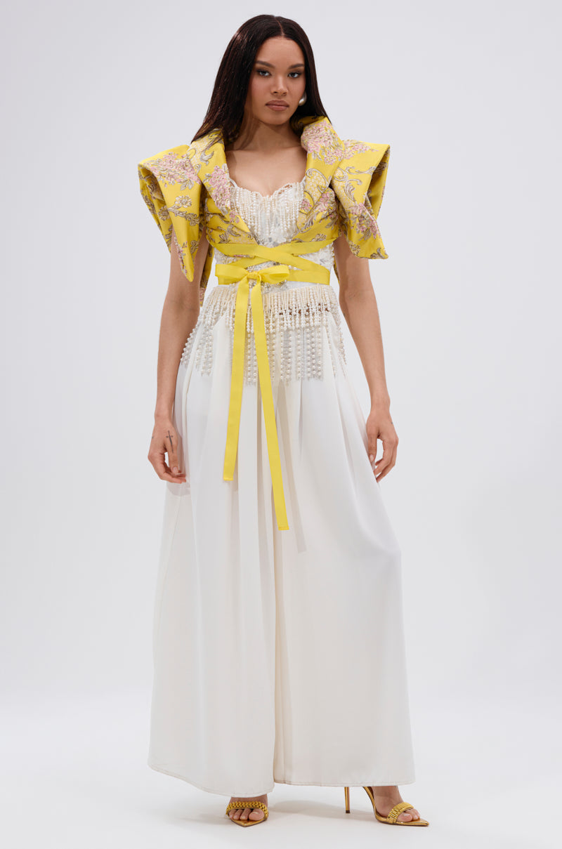 A woman wears the DION STATEMENT BOLERO IN YELLOW with a yellow floral jacquard design, sculptural shoulders, lace-trimmed bodice, yellow waist sash, and pairs it with flowing white wide-leg pants and heeled sandals. She has long straight hair.