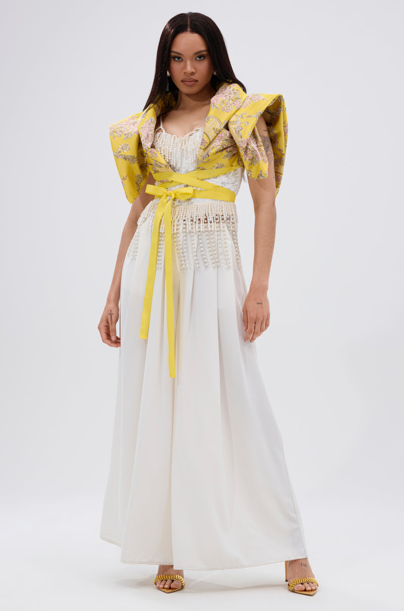 A woman wears the DION STATEMENT BOLERO IN YELLOW, a yellow floral jacquard bolero with sculptural shoulders, over a white floor-length dress with a fitted textured bodice, accessorized with a yellow sash and open-toe yellow shoes.