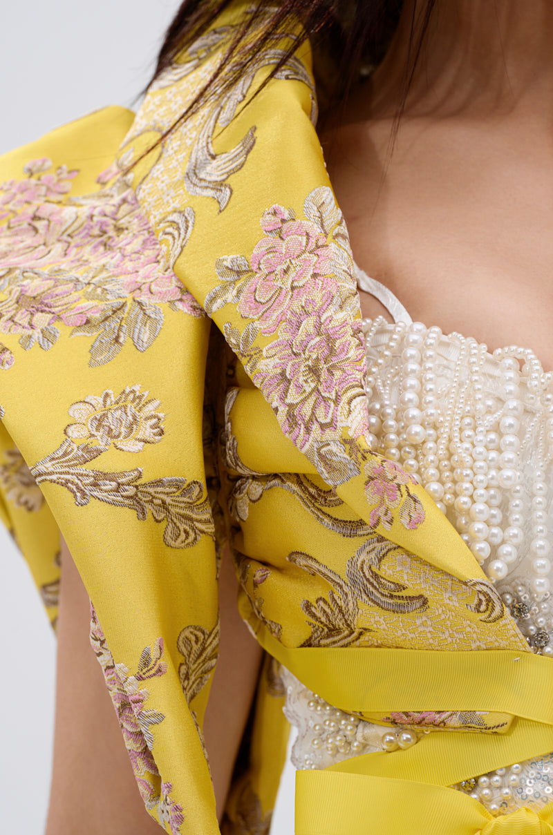 A close-up of a woman wearing the DION STATEMENT BOLERO IN YELLOW, featuring pink floral embroidery and a pearl-embellished bodice, with fabric elegantly draped over her sculptural shoulders and chest.
