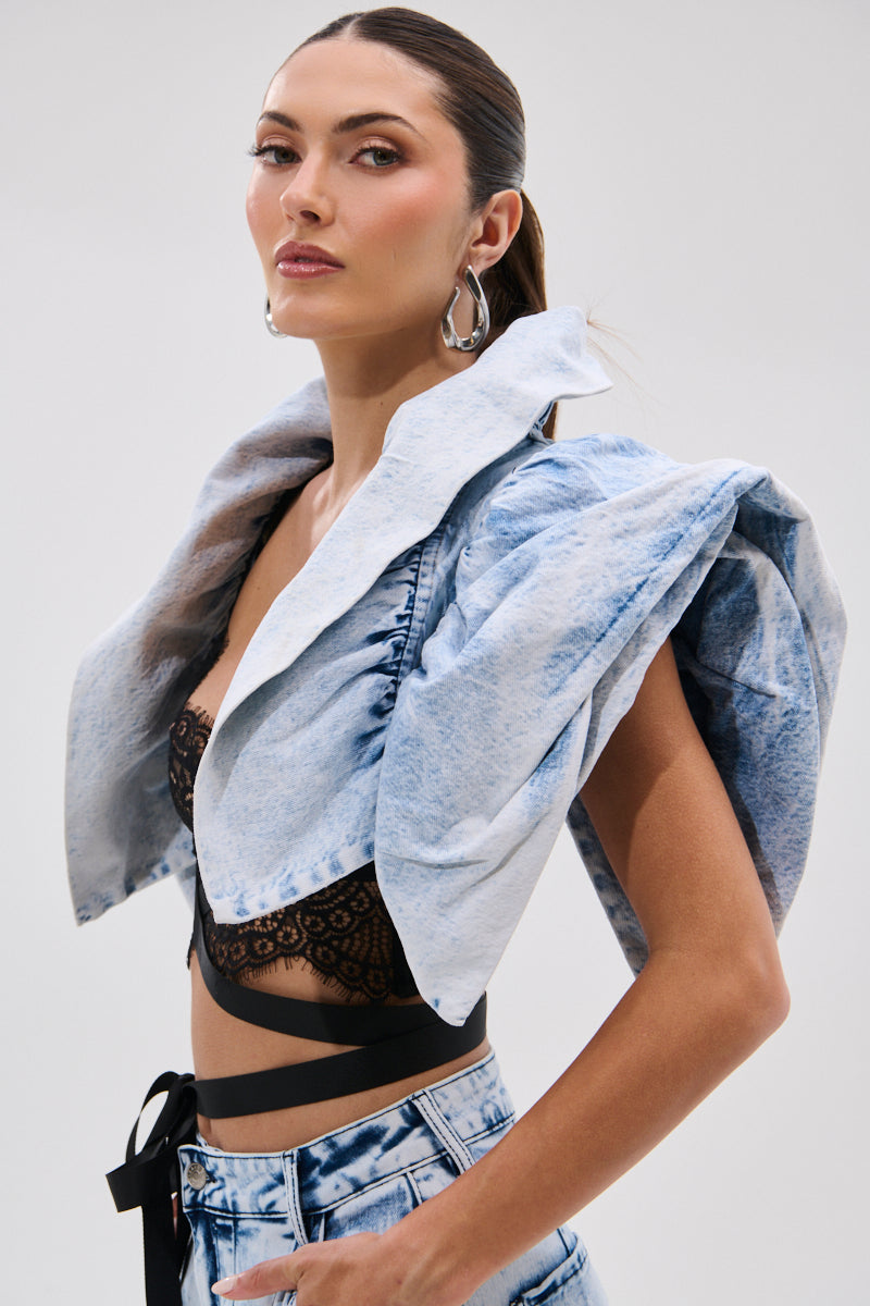 A woman poses in the DION BOLERO IN DENIM, a light blue sculpted jacket with exaggerated shoulders, styled with a black lace crop top, high-waisted acid-wash jeans, large hoop earrings, and slicked-back hair.