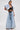 A woman models the DION BOLERO IN DENIM with large puff sleeves, paired with wide-leg, light-wash jeans and a cropped black lace top. She completes the look with hoop earrings and strappy sandals against a plain backdrop.