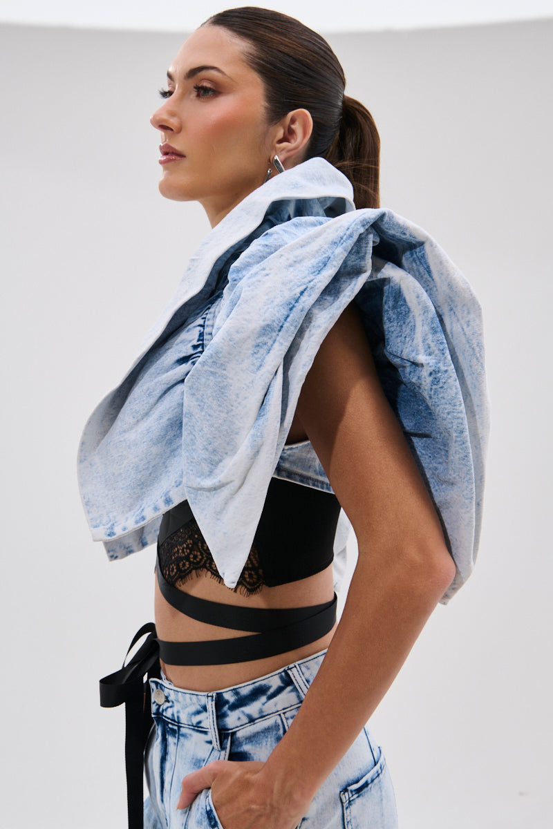 A woman in profile wears the DION BOLERO IN DENIM draped over one shoulder with a black crop top and high-waisted acid-wash jeans. Her hair is pulled back, set against a plain white background.
