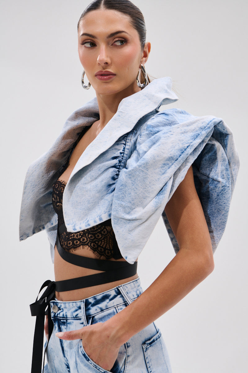A woman wears the DION BOLERO IN DENIM, a light blue cropped jacket with bold shoulders, over a black lace bralette and high-waisted, light wash jeans. She completes the look with slicked-back hair, hoop earrings, and a confident expression.