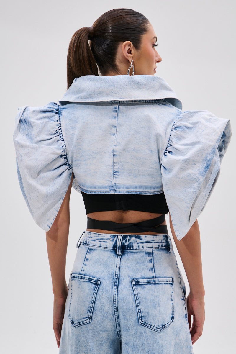 A woman with a ponytail, seen from the back against a light gray background, wears the DION BOLERO IN DENIM with puff sleeves and matching high-waisted jeans.