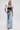 A woman stands in a studio wearing the DION BOLERO IN DENIM over a black lace crop top, paired with high-waisted wide-leg acid-wash jeans. She completes the look with hoop earrings, a ponytail, and strappy heels.