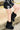 A person in a black skirt with white lace-up details pairs their look with AZALEA WANG DIPA BLACK FUR FLAT BOOTIE, standing outdoors on concrete and leaning against a yellow railing.