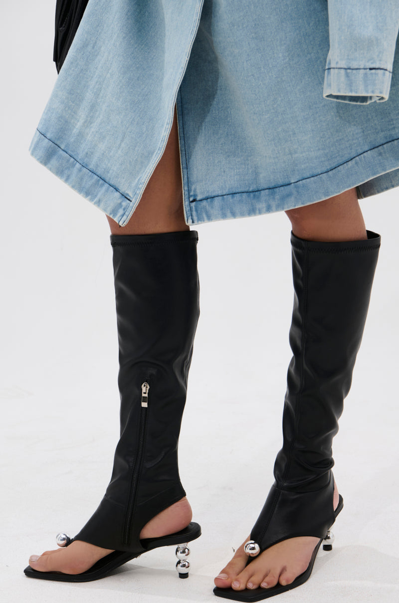 A person wears a light blue denim skirt and AZALEA WANG DIPPIN DOT BLACK STRETCH SANDAL BOOTS—unique faux leather knee-highs with open square toes, zippers, and metallic ball accents on flat soles.