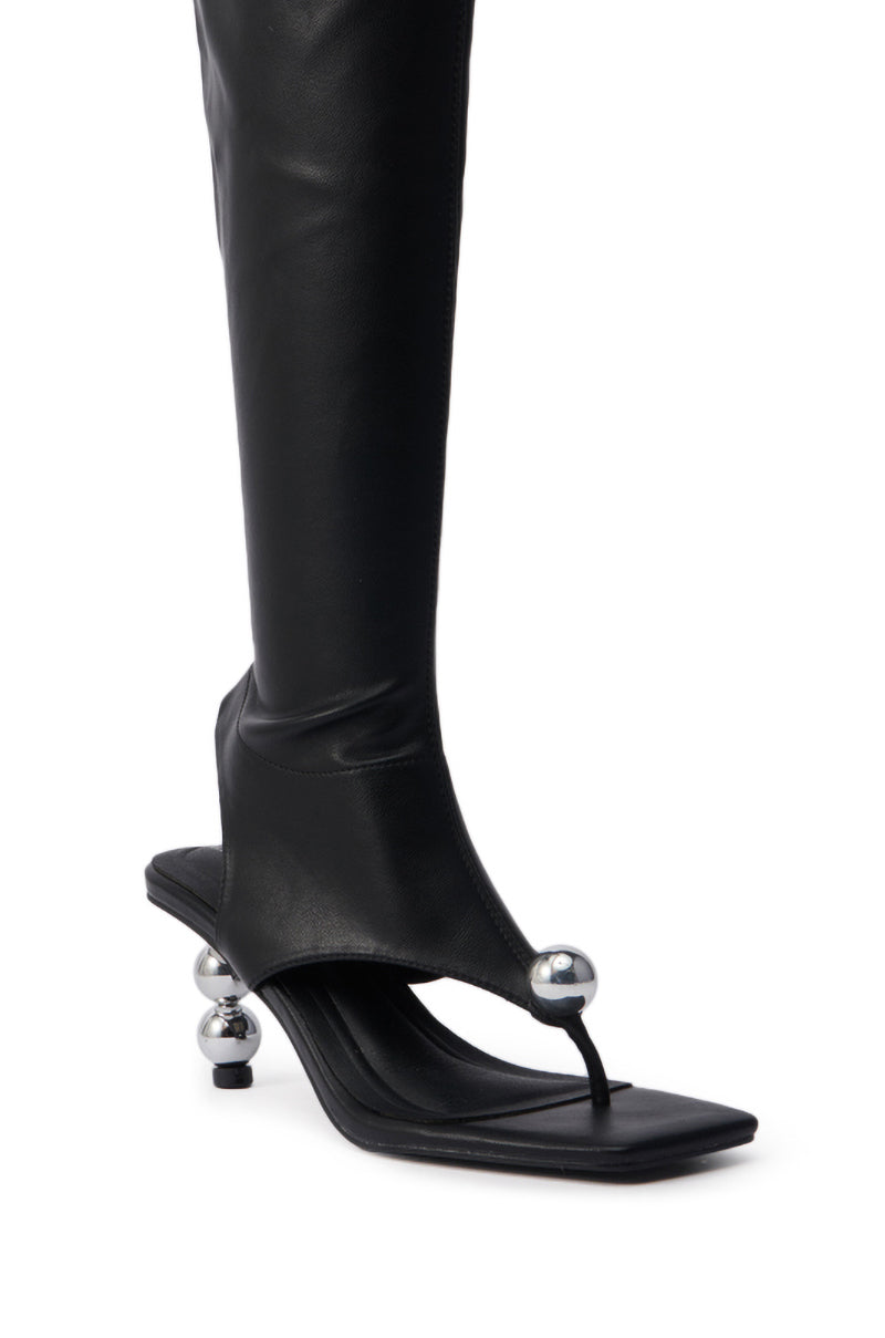 A close-up of the AZALEA WANG DIPPIN DOT BLACK STRETCH SANDAL BOOT—black knee-high with an open square toe, metallic silver ball accents on the front strap and heel—styled with black, fitted thigh-high stockings.