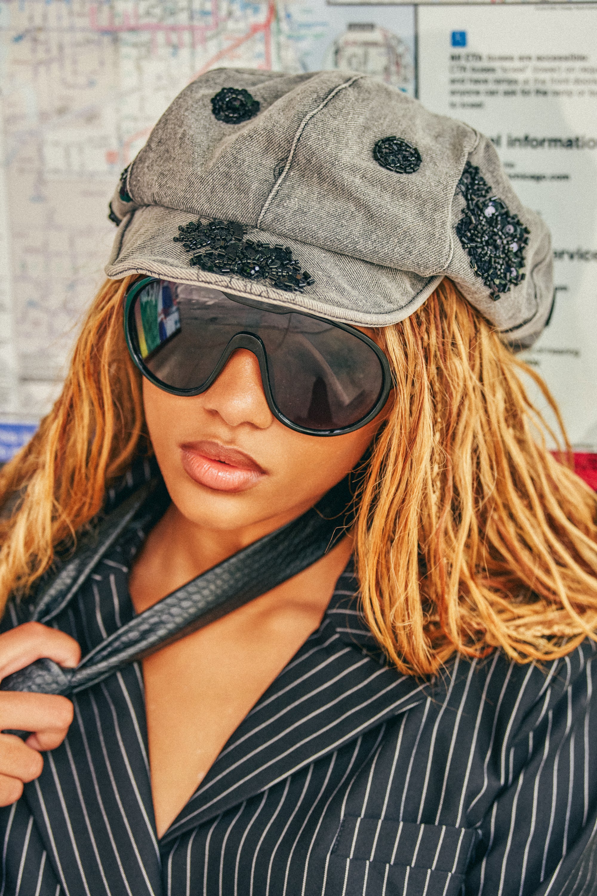 Wearing the Y2K DREAMS CABBIE HAT in grey denim, a person with blond dreadlocks, large dark sunglasses, and a pinstripe blazer featuring subtle sequin embroidery holds a black strap while standing in front of a transit map.