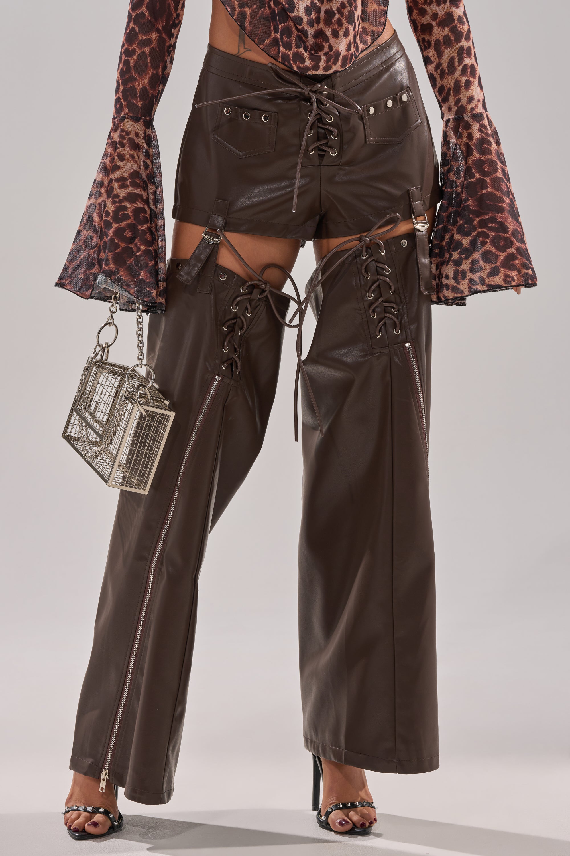 A person styles the WANNA BE FAUX LEATHER CHAPS with detachable sections over western shorts, pairs them with a leopard print flared sleeve top, and finishes the festival look with a silver cage handbag.