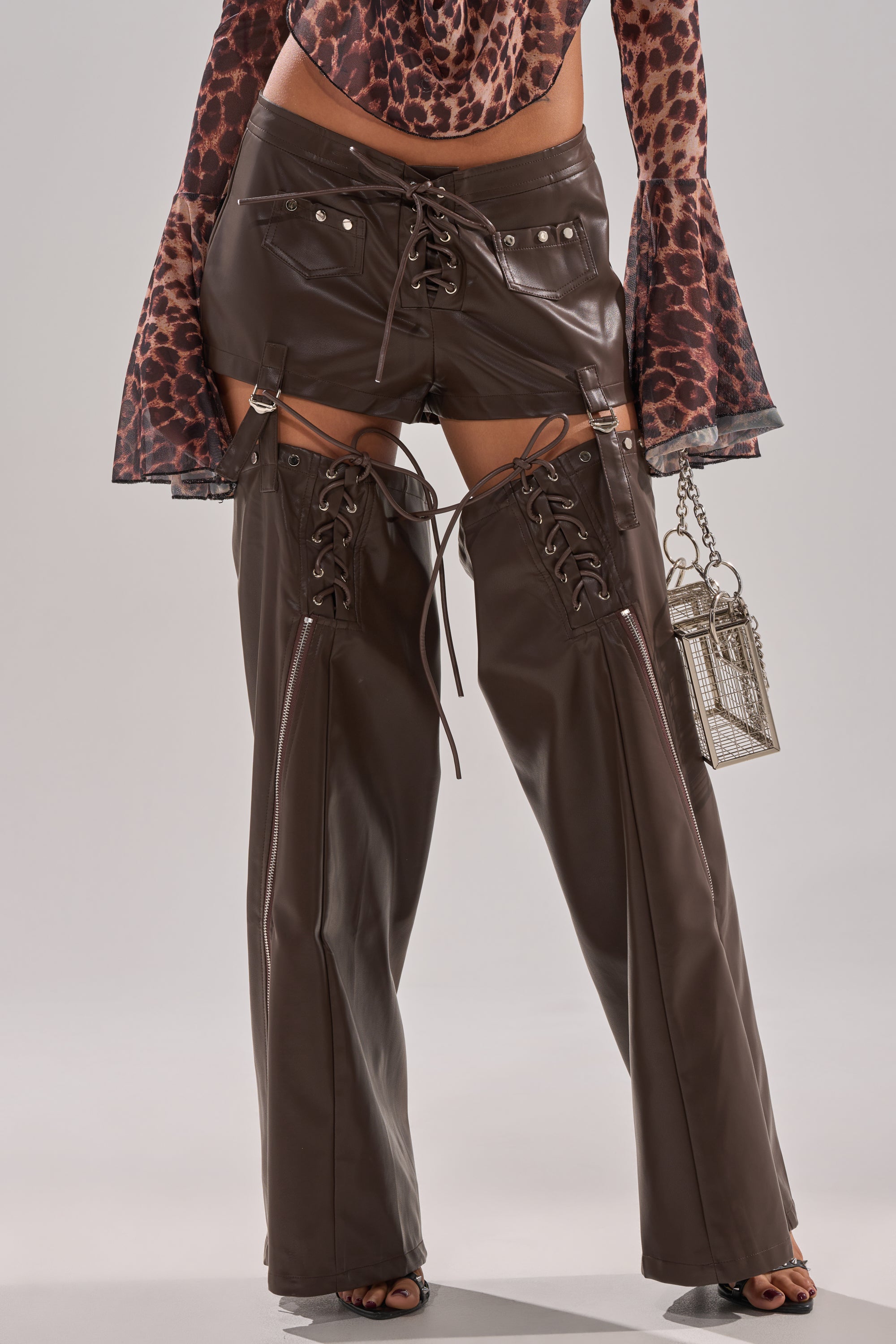 A person in WANNA BE FAUX LEATHER CHAPS—high-waisted, dark brown with lace-up details and detachable thighs—pairs them with a leopard print long-sleeve top and a geometric metallic handbag for a bold festival look.
